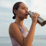 A woman drinks from a metal bottle on a sunny day by the beach, wearing a sports bra.